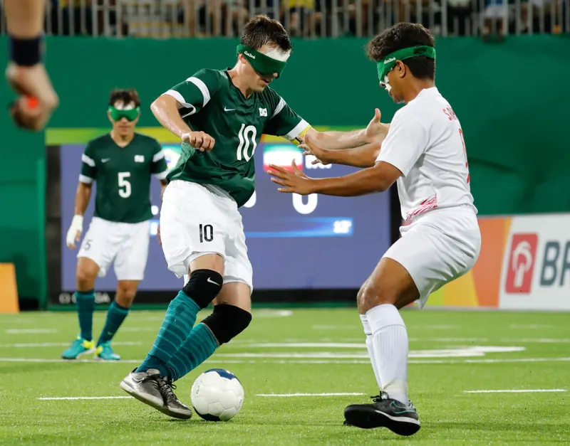 Ricardinho, de camisa verde número 10 e venda nos olhos, conduz a bola no gramado. Um adversário de branco estende os braços para alcançá-la; ao fundo, um companheiro de camisa 5.