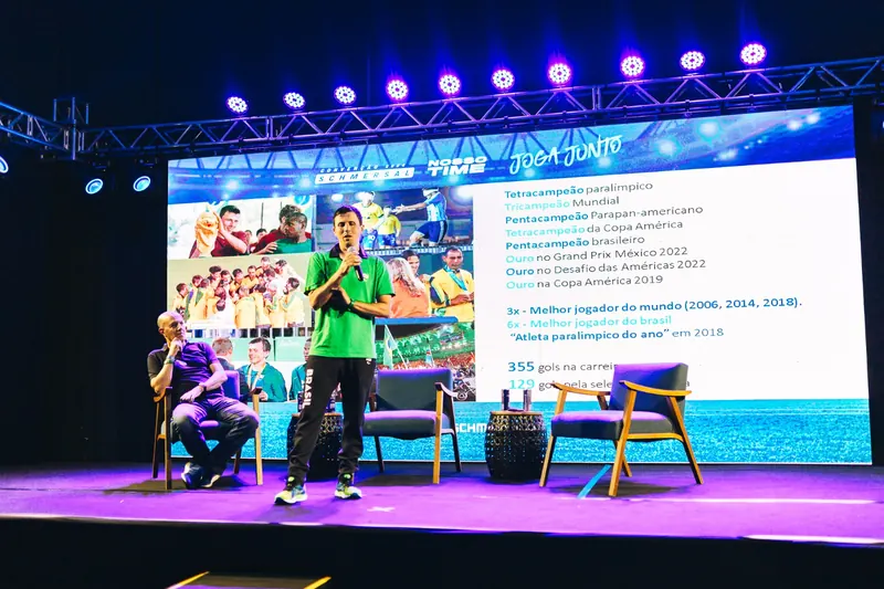 Ricardinho de pé no palco, de camisa verde e calça da Seleção Brasileira, fala ao microfone. Ao fundo, um telão apresenta sua carreira; ao lado, um participante está sentado.