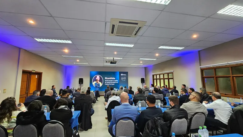 Auditório lotado, visto de fundo, com pessoas sentadas em mesas azuis. Ao fundo, Ricardinho apresenta diante de uma tela com o nome dele.